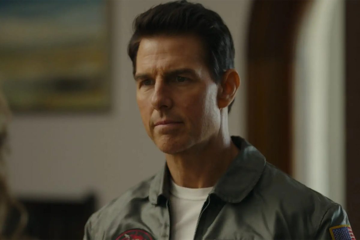 A man stands calmly inside, wearing a military-style jacket, with a blurred painting and neutral walls in the background.
