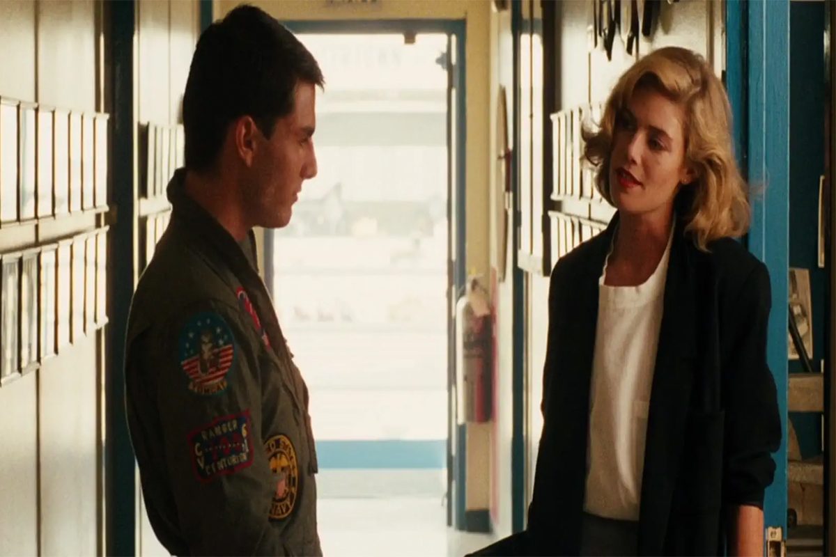 Man and woman talking in a corridor, with the man wearing a military flight jacket adorned with patches; background features open doors and hallway.