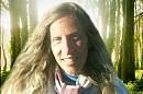 A person smiles gently, standing amidst a forest with sunlight filtering through tall trees, creating a serene and natural atmosphere.
