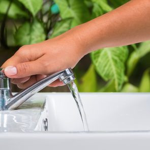 A hand turns on a shiny faucet, releasing water into a white sink; green leaves are visible in the background.