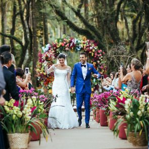 Couple walks joyfully down aisle; guests photograph surrounded by colorful flowers in outdoor wedding setting.