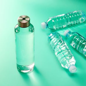 A glass water bottle stands upright; nearby, four plastic bottles lie scattered on a turquoise surface.