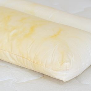 Yellowed pillow lies on a mattress, displaying noticeable stains, within a bedroom setting.