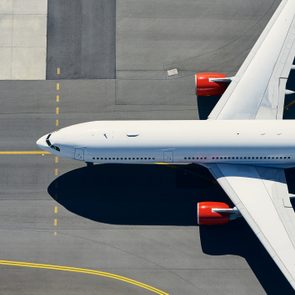 Airplane taxis on the runway, viewed from above, featuring distinct red engines and white fuselage against a gray tarmac background with yellow guide lines.
