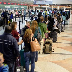 People stand in line with luggage at an airport check-in counter, surrounded by signs and digital screens.