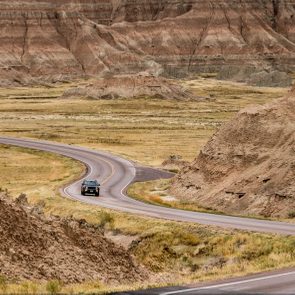 A car drives along a winding road, surrounded by rugged, eroded landscapes and grassy plains.