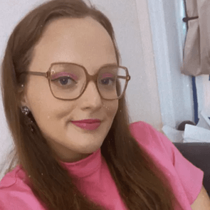 Person smiling, wearing glasses and a pink top, in a casual indoor setting with white walls and some visible furniture.