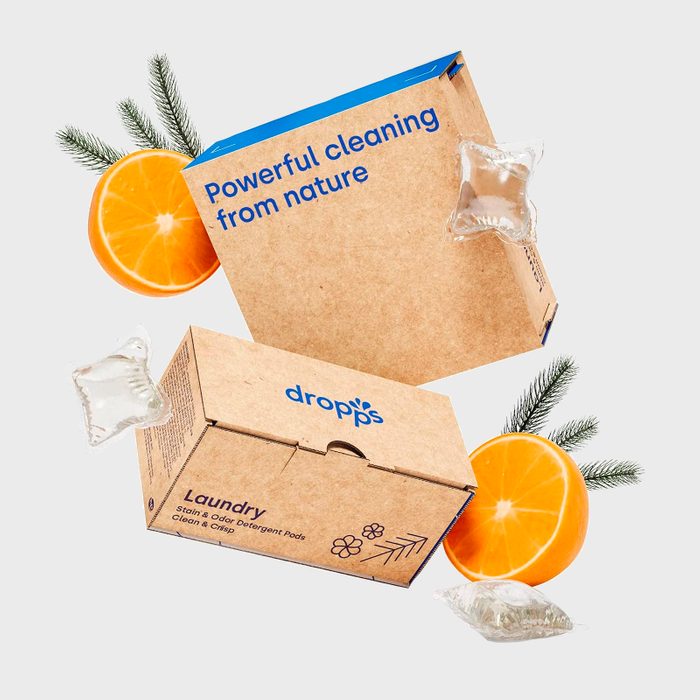 Boxes display text promoting natural cleaning, surrounded by detergent pods, half an orange, and pine sprigs on a light background.