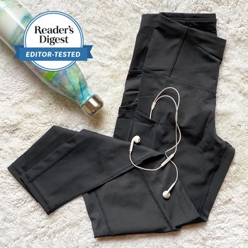 Black leggings lie on a textured surface with white earbuds placed on them. A patterned water bottle is nearby. Text: "Reader's Digest EDITOR-TESTED."