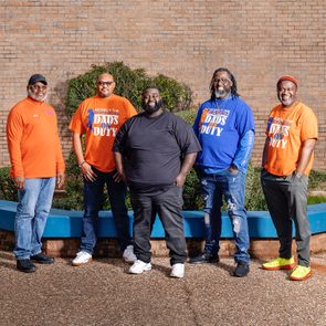 Five men stand, smiling, against a brick wall. Three wear 