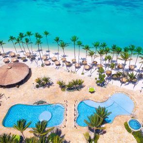 Pools shimmer by palm tree-lined beachfront, surrounded by sun loungers and thatched umbrellas, adjacent to a circular, open-air pavilion.