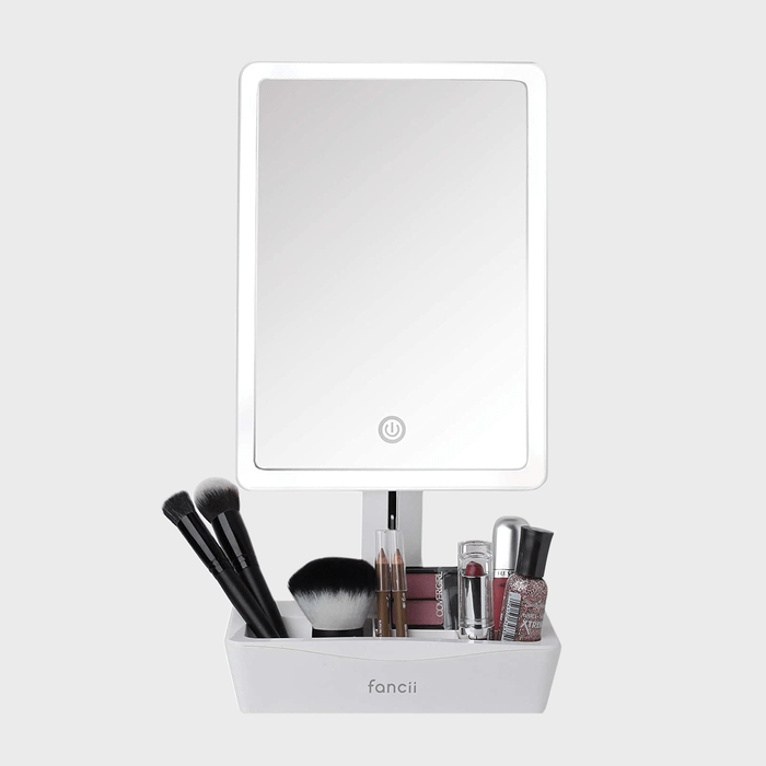 Mirror displays power button, standing on a base. Surrounding are makeup brushes and cosmetics, neatly organized in a white tray marked 