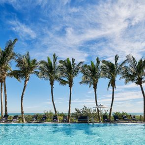 Pool reflects palm trees, sun loungers, and umbrellas in a beachside setting under a bright blue sky.