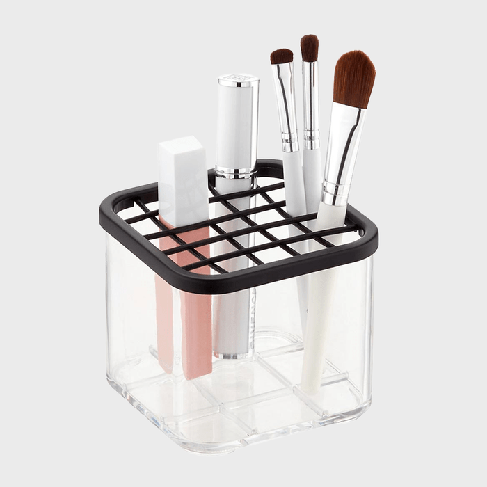 A clear container holds makeup brushes and lipsticks upright, organized by a black grid, against a plain background.