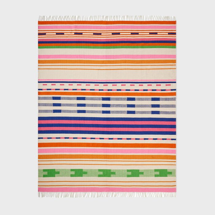 A colorful, striped rug lies flat, featuring vibrant horizontal patterns in orange, blue, green, and pink, with fringed edges against a plain background.