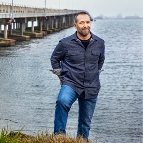 Man stands smiling on grass beside water, bridge extends in the background under cloudy sky.