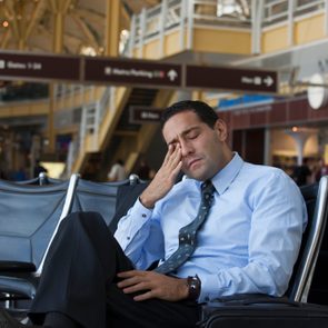 Man rests in airport chair, appearing tired, with closed eyes and hand on face; signage above indicates gate directions and parking.