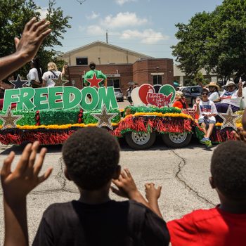 Float displays "FREEDOM," people wave in a colorful parade, children waving in foreground; sunny day, trees and buildings in background.