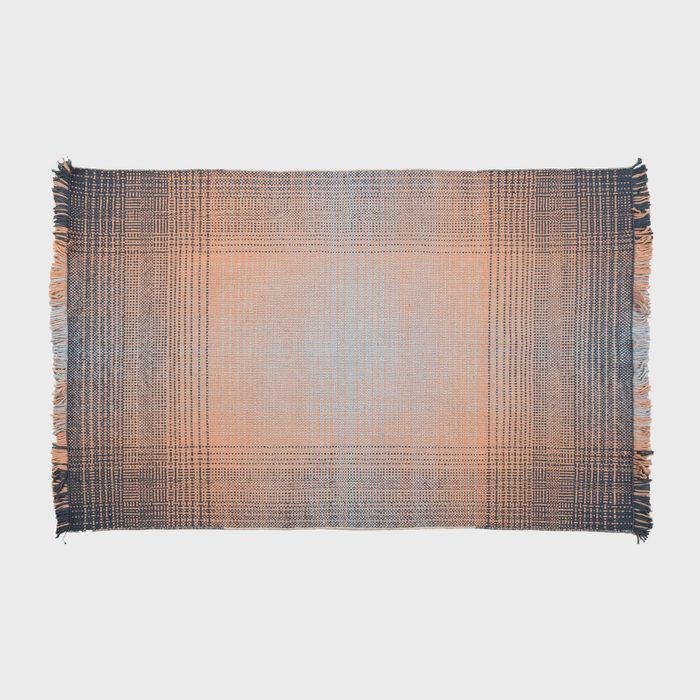 Woven rug displays intricate plaid pattern with fringed edges, lying flat on a plain background.