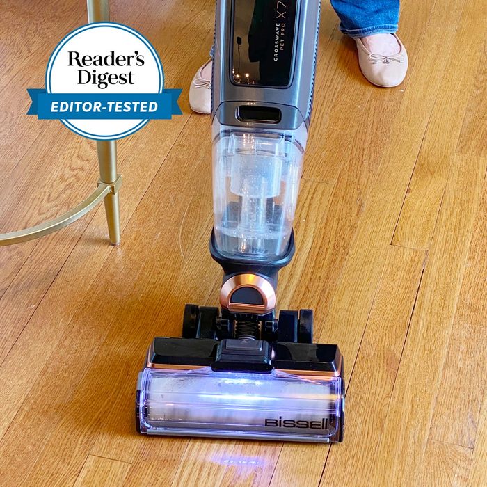 Vacuum cleaner cleans wooden floor; white shoes and table leg visible nearby. 