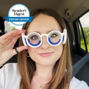 Woman wears circular white and blue glasses, adjusts them in a car. Badge reads 