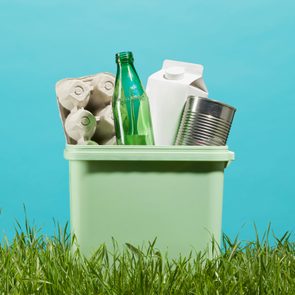 A recycling bin containing paper, glass, carton, and metal sits on grass against a blue background.