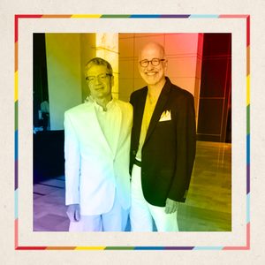 Two men stand and smile indoors, both wearing blazers and glasses. The image features rainbow tinting and frame, creating a colorful effect.