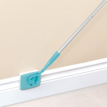 Mop scrubs white baseboard; turquoise cleaning head and handle, beige wall background.