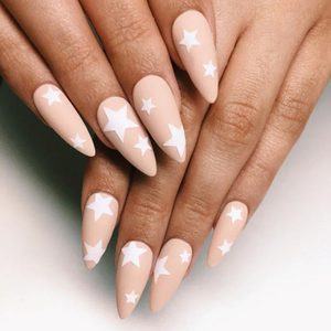 Hand with pointed nails displays white star designs on a nude background, creating a celestial-themed manicure.