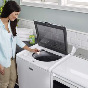 A woman places a tablet into a top-load washing machine in a laundry room, with folded towels and cleaning product nearby.