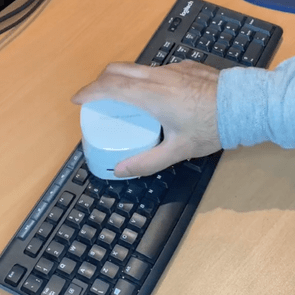Hand cleans keyboard with small, white cleaning device on wooden desk.