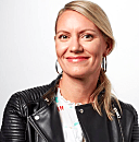 A person stands smiling, wearing a black leather jacket, in front of a plain white background.