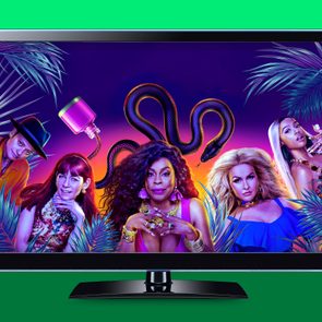 Women pose confidently in vibrant tropical-themed setting with palm leaves and large snake, displayed on a television screen in a green background.