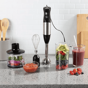 Blender apparatus stands on countertop; surrounded by fruits, vegetables, juice, and cooking utensils; kitchen setting with white tiled wall and gray countertop.