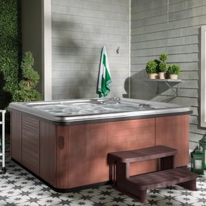 Hot tub with bubbling water beside a small wooden step, situated on patterned tiles near greenery and a wall with potted plants.