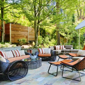 Patio furniture arranged under a large tree with woven chairs, orange cushions, and striped pillows in a garden with wooden fence and greenery.