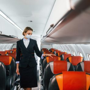 Flight attendant walking down airplane aisle, wearing a mask, among empty seats with red and orange headrests.
