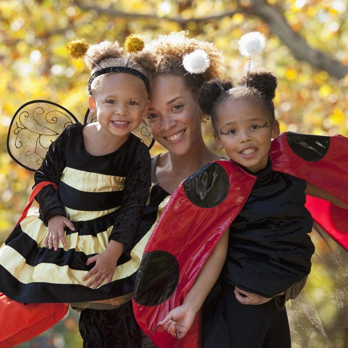 Two children in bee and ladybug costumes smile, held closely by a smiling adult in a sunlit, autumnal park setting.