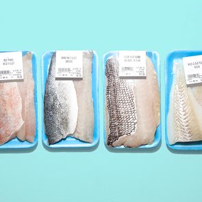 Four packages of fish fillets displayed on a light blue surface, labeled with type, weight, price, and expiry date on white stickers.