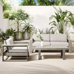 Outdoor furniture set with a sofa, chair, and table; features gray cushions and wooden frames, surrounded by lush tropical plants on a wooden deck.