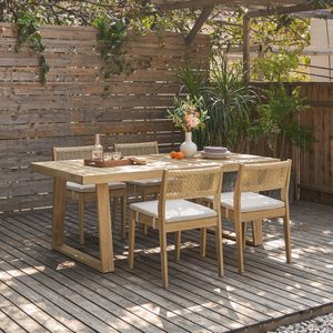 Wooden dining table displays vase, drinks, and fruits, surrounded by chairs on a patio. Wooden fence and lush plants create a serene atmosphere.