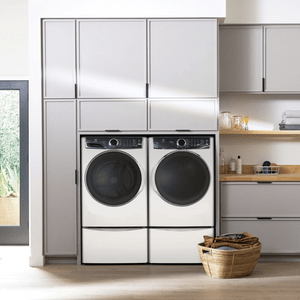 Front-loading washers sit idle in modern laundry room; nearby shelves hold towels, bottles, and a basket.
