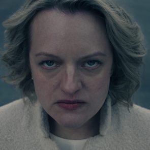A person with a serious expression looks directly at the camera, wearing a light-colored coat against a blurred, dark background.