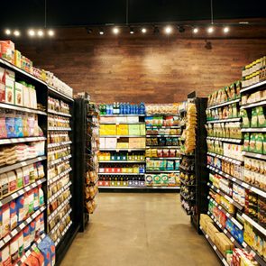 Shelves display various packaged goods in a grocery store aisle, under warm lighting with a wooden wall backdrop.