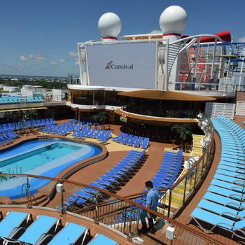 Deck chairs surround a rectangular pool on a cruise ship under a clear blue sky, with people strolling nearby. Carnival logo visible on screen.