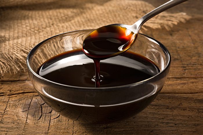 Spoon drips dark liquid into glass bowl on rustic wooden surface with burlap fabric nearby.