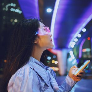 Person wearing neon glasses holds a smartphone, looking upwards under purple streetlights in a city setting.