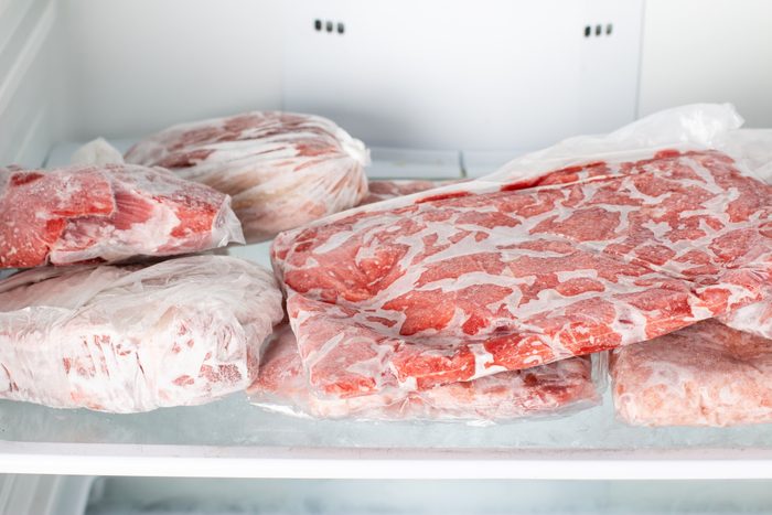 Frozen meat wrapped in plastic lies stacked on a white refrigerator shelf.