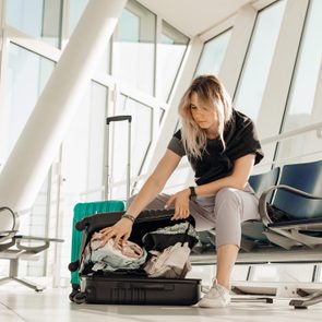 A woman organizes clothes in a suitcase at an airport, seated between modern chairs beneath large windows.