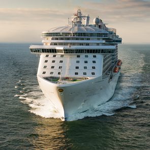 Cruise ship sailing forward, cutting through ocean waters, under a partly cloudy sky.